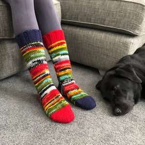 Op de afbeelding: Een paar kleurrijke gestreepte gebreide sokken met rode, gele, blauwe, groene en witte strepen. De sokken worden gedragen door een persoon die op een bank zit met een zwarte hond die op de vloer slaapt.