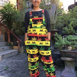 May include: A person wearing black and green overalls with a marijuana leaf and lion pattern. The overalls have adjustable straps and are made of a lightweight fabric.