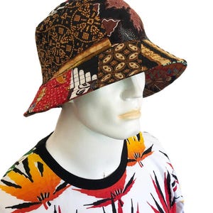 Op de afbeelding: Een zwarte bucket hat met een patchwork ontwerp met verschillende patronen in tinten bruin, rood en wit. De hoed is gemaakt van katoenen stof en heeft een brede rand.