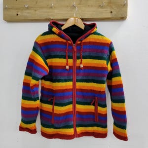 Könnte beinhalten: Ein bunter, gestreifter Kapuzenpullover mit einem roten Reißverschluss. Der Pullover hat ein Regenbogenmuster mit Streifen in Rot, Orange, Gelb, Grün, Blau, Lila und Grau.