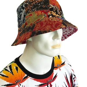 Op de afbeelding: Een veelkleurige bucket hat met een patchwork ontwerp. De hoed is gemaakt van katoenen stof en heeft verschillende patronen, waaronder bloemen, geometrische vormen en paisley. De hoed is omkeerbaar, met een effen rode kant en een gepatroneerde kant.