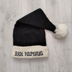 Puede incluir: Gorro de Papá Noel negro tejido con un borde blanco. El borde presenta la frase bordada "BAH HUMBUG" en negro. El gorro tiene una forma larga y puntiaguda y un pompón blanco en el extremo.