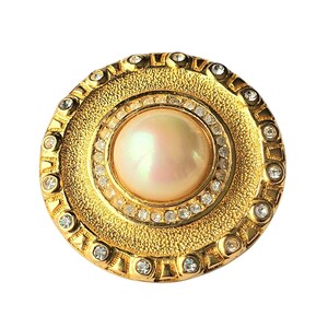 May include: A gold-toned, round brooch with a large, iridescent pearl center. The brooch features a textured outer ring, a ring of clear crystals, and a decorative border. The design is symmetrical and elegant, suitable for formal wear.