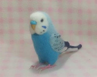 Puede incluir: Un pájaro de fieltro azul y blanco con un pico azul claro y ojos negros. El pájaro está sentado sobre una superficie con estampado rosa y blanco.