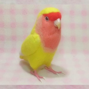 May include: A small, yellow and pink stuffed bird with a red beak. The bird is made of felt and has a fluffy texture.