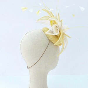 May include: A yellow and white straw hat with a large, decorative bow and feathers. The hat is on a white mannequin head.