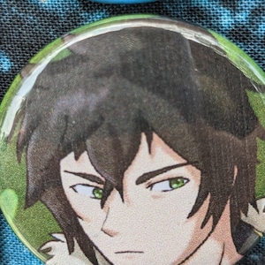 May include: A close-up of a button featuring a cartoon character with dark green hair, green eyes, and a serious expression. The character is wearing a light brown collar with a gold trim.