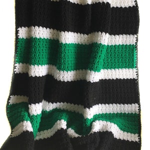 May include: A striped crocheted blanket in black, white, and green. The blanket is folded and draped over a white surface.