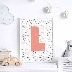 May include: A white framed print with a pink letter L on a white background with black hearts. The name Lily is written below the letter.