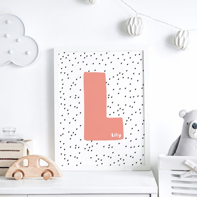 May include: A white framed print with a pink letter L on a white background with black hearts. The name Lily is written below the letter.