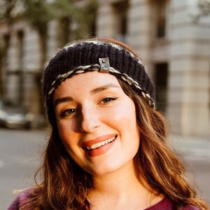 May include: A woman with long brown hair is wearing a purple sweater and a black and white knit headband. She is smiling and looking at the camera. The background is a city street with buildings and cars.