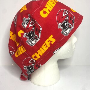 May include: A red fabric cap featuring a repeating pattern of yellow text that reads "CHIEFS" and illustrations of football helmets. The cap is designed to fit the head and has a tie closure.