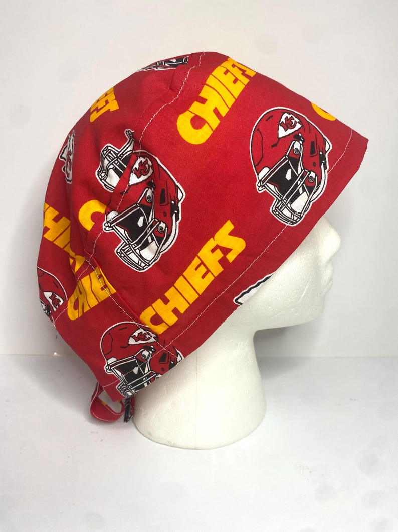 May include: A red fabric cap featuring a repeating pattern of yellow text that reads "CHIEFS" and illustrations of football helmets. The cap is designed to fit the head and has a tie closure.