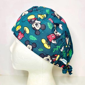 May include: Teal scrub cap featuring a repeating pattern of Mickey Mouse in various poses, with yellow, red, and white accents. Green leaves are interspersed throughout the design. The cap is tied at the back.