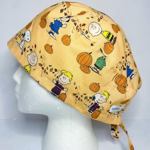 May include: A light yellow scrub cap featuring a repeating pattern of cartoon characters, pumpkins, and falling leaves. The characters include Snoopy, Charlie Brown, and other Peanuts characters. The cap has a tie in the back.