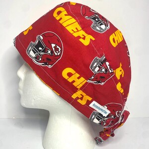 May include: A red and yellow Chiefs print scrub cap. The cap features the word "CHIEFS" in yellow, along with football helmets. The cap is on a white mannequin head. The tag says "RainydayWaffles".