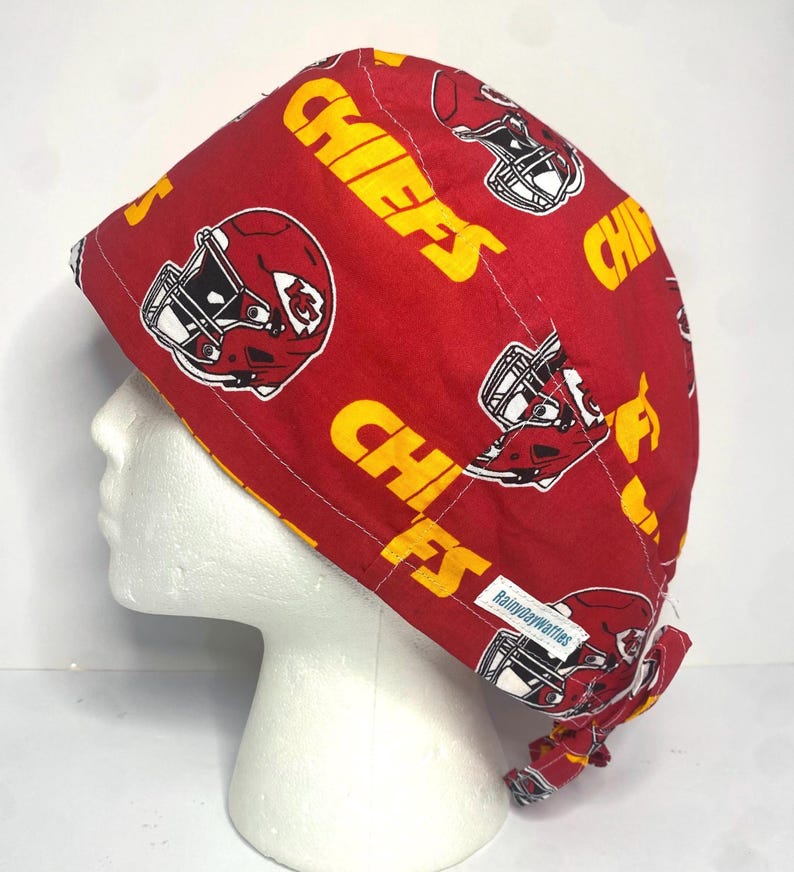 May include: A red and yellow Chiefs print scrub cap. The cap features the word "CHIEFS" in yellow, along with football helmets. The cap is on a white mannequin head. The tag says "RainydayWaffles".