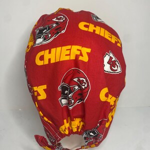 May include: Red and gold Chiefs themed scrub cap. The cap features repeating patterns of the team's helmet and logo, along with the word "CHIEFS" in gold lettering. The cap has a tie in the back.
