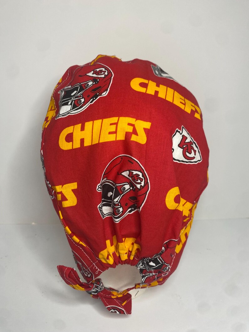 May include: Red and gold Chiefs themed scrub cap. The cap features repeating patterns of the team's helmet and logo, along with the word "CHIEFS" in gold lettering. The cap has a tie in the back.