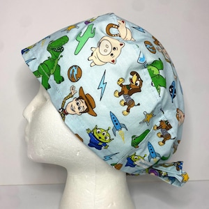 May include: A light blue surgical scrub cap featuring a pattern of cartoon characters, including a green dinosaur, a cowboy, and an alien. The cap is tied at the back and is designed to cover the hair during medical procedures.