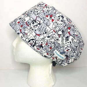 May include: A white surgical scrub cap featuring a repeating pattern of various dog breeds in gray, black, and red. The cap has a drawstring closure and a small tag that reads "RainydayWarries".