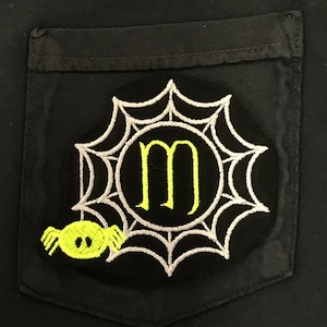 Puede incluir: Un bolsillo de tela negro con un diseño de telaraña bordado en amarillo y blanco. La telaraña tiene una letra "M" amarilla en el centro y una araña amarilla colgando de la parte inferior.