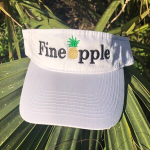 May include: White visor with a black embroidered pineapple and the word "Fine" on the left and "apple" on the right.