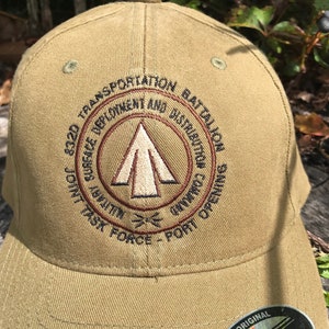 May include: A tan baseball cap with an embroidered logo that reads "322D TRANSPORTATION BATTALION JOINT TASK FORCE - PORT OPENING SURFACE DEPLOYMENT AND DISTRIBUTION COMMAND". The logo features a brown tent with a white interior and a brown arrow pointing upwards. The cap has a black Flexfit label on the back.