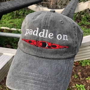 May include: A gray baseball cap with the text "paddle on" embroidered in white. A red kayak with black details is embroidered below the text.