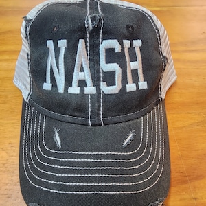 May include: A black and white trucker hat with a distressed look. The hat has a white mesh back and a black front with the word "NASH" embroidered in white.