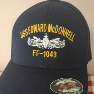 May include: A navy blue baseball cap with the embroidered text "USS EDWARD MCDONNELL FF-1043" in yellow and white. The cap has a Flexfit label on the side.