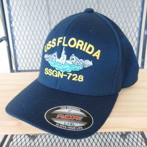 May include: A navy blue baseball cap with the text "USS FLORIDA SSGN-728" embroidered on the front. The cap features a yellow outline and a small image of a submarine. The cap is labeled "Flexfit" and "L/XL".