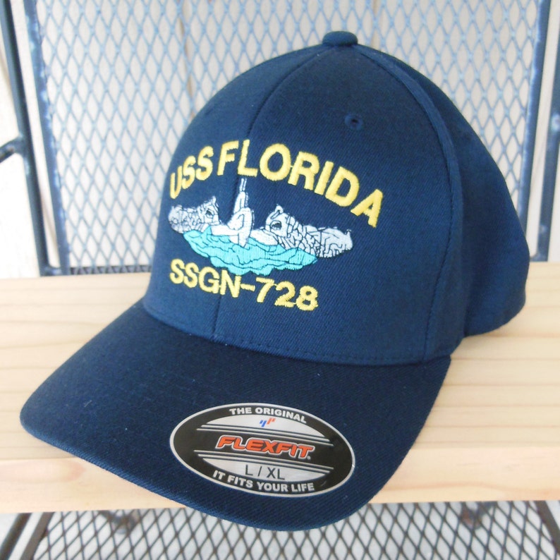 May include: A navy blue baseball cap with the text "USS FLORIDA SSGN-728" embroidered on the front. The cap features a yellow outline and a small image of a submarine. The cap is labeled "Flexfit" and "L/XL".