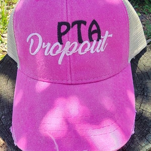 May include: A pink trucker hat with a distressed brim and a beige mesh back. The hat features the embroidered text "PTA Dropout" in black and white script.