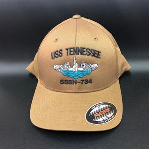May include: A tan Flexfit baseball cap with the text "USS TENNESSEE" embroidered above a blue and white graphic of a submarine. The text "SSBN-734" is below the graphic. The hat has a curved brim and a Flexfit sticker.