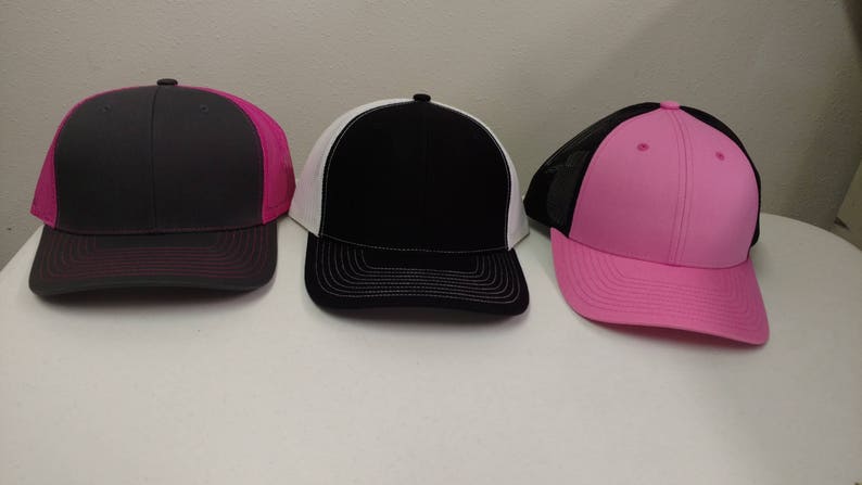 May include: Three trucker hats in different colors. The first hat is gray with a pink mesh back. The second hat is black with a white mesh back. The third hat is pink with a black mesh back.