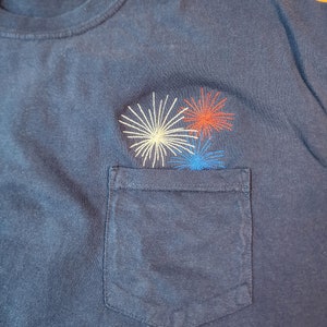 May include: A navy blue t-shirt with a pocket featuring an embroidered design of three fireworks in red, white, and blue.