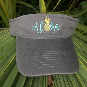 May include: A black visor with a pineapple and the word "Aloha" embroidered in yellow and turquoise.