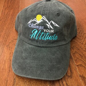 K&ouml;nnte beinhalten: Eine dunkelgraue Baseballkappe mit einem gestickten Design aus Bergen, einer Sonne und dem Text "Change Your Altitude".