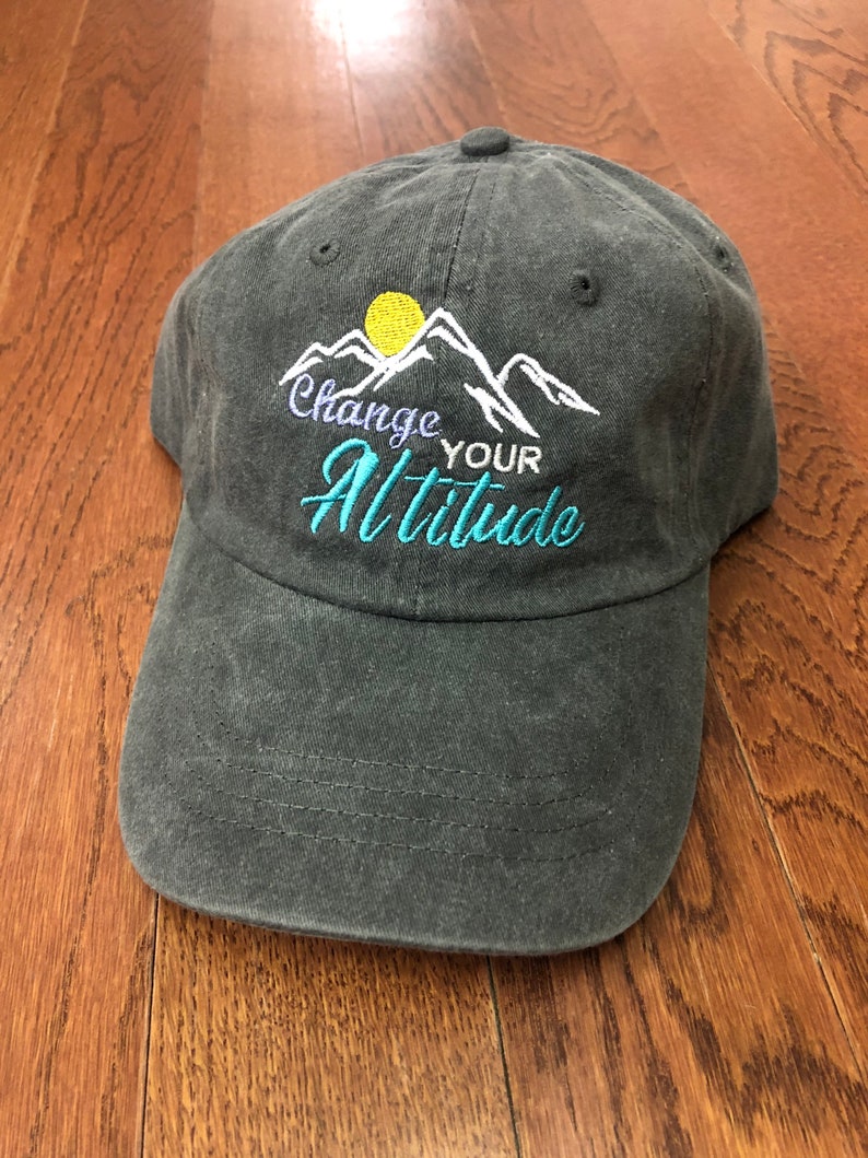 K&ouml;nnte beinhalten: Eine dunkelgraue Baseballkappe mit einem gestickten Design aus Bergen, einer Sonne und dem Text "Change Your Altitude".