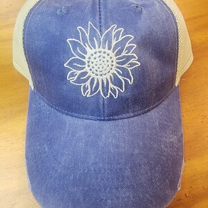 May include: A blue and tan distressed trucker hat with a white embroidered sunflower design.