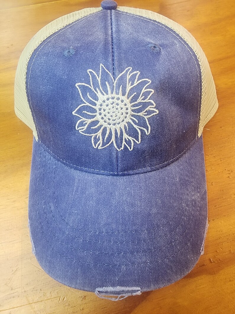 May include: A blue and tan distressed trucker hat with a white embroidered sunflower design.