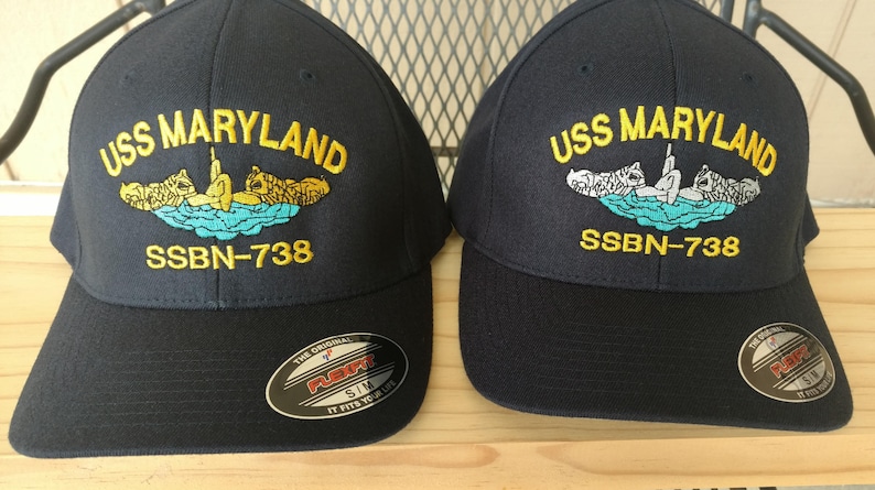 May include: Two black baseball caps with embroidered navy blue patches. The patches feature the text "USS MARYLAND" and "SSBN-738" with a yellow submarine graphic.