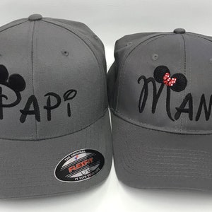 May include: Two gray baseball caps. One cap has the word "Papi" embroidered in black, with a Mickey Mouse-inspired design. The other cap has the word "Mami" embroidered in black, with a Minnie Mouse-inspired design, including a red bow.