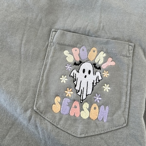May include: A gray t-shirt with a pocket featuring an embroidered design of a ghost with the words "Spooky Season" in colorful letters.