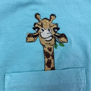 May include: A light blue t-shirt with a brown and white embroidered giraffe design. The giraffe is looking at the viewer and has its head sticking out of a pocket on the shirt.