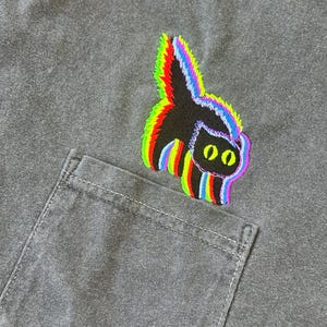 May include: A black cat with rainbow-colored outlines is embroidered on a gray t-shirt. The cat is looking to the right and has yellow eyes.
