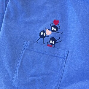May include: A blue t-shirt with a pocket and an embroidered design of three black and white creatures holding a red heart.