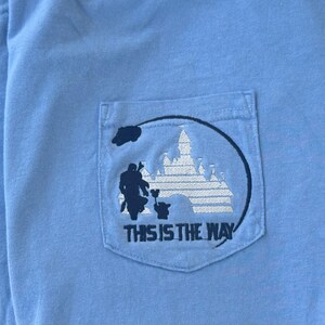 May include: A light blue t-shirt with a pocket featuring an embroidered design of a Mandalorian character with a Grogu character, a castle, and the text "This is the Way".