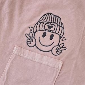 May include: A pink t-shirt with a black embroidered smiley face wearing a beanie with a heart on it. The smiley face is making a peace sign with both hands.